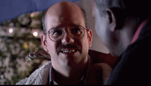 A Bald Man With Glasses And A Mustache Smiles While Talking To Another Man GIF