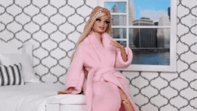A Barbie Doll Wearing A Pink Robe Is Sitting On A Bed In Front Of A Window . GIF
