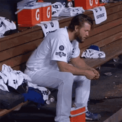 A Baseball Player Is Sitting On A Bench In A Dugout With Gatorade Coolers In The Background GIF