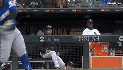 A Baseball Player Is Swinging At A Ball While A Man Sits In The Dugout Watching . GIF