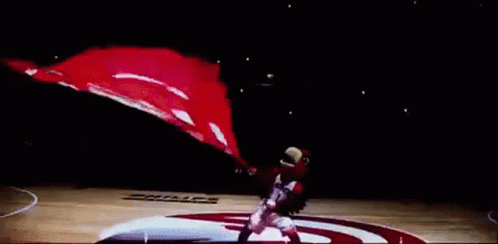 A Basketball Player Is Holding A Basketball In Front Of A Large Hawks Flag GIF