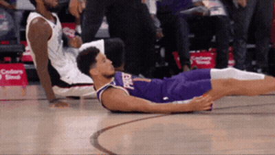 A Basketball Player Is Laying On The Floor While Another Player Sits On The Bench GIF
