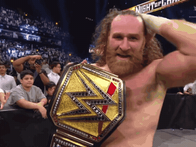 A Bearded Wrestler Is Holding A Wrestling Championship Belt In Front Of A Night Of Champions Sign GIF