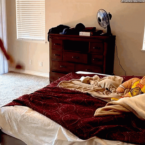 A Bedroom With A Fan On The Dresser And A Bed With A Red Blanket GIF