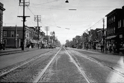 A Black And White Photo Of A City Street With A Train Going Down It GIF