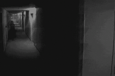 A Black And White Photo Of A Dark Hallway With A Person Walking Down It GIF