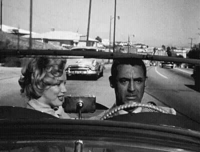 A Black And White Photo Of A Man And A Woman Driving A Car Down A Street . GIF