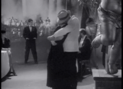 A Black And White Photo Of A Man And Woman Hugging In Front Of A Crowd GIF