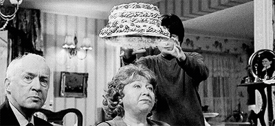 A Black And White Photo Of A Man Holding A Cake Over A Woman 'S Head In A Living Room . GIF