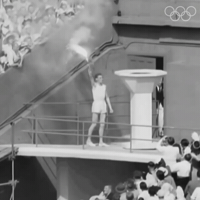 A Black And White Photo Of A Man Holding A Torch With The Olympic Rings On The Wall Behind Him GIF