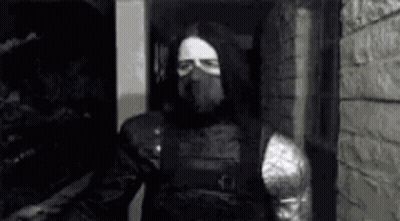 A Black And White Photo Of A Man In A Mask Standing In Front Of A Brick Wall . GIF