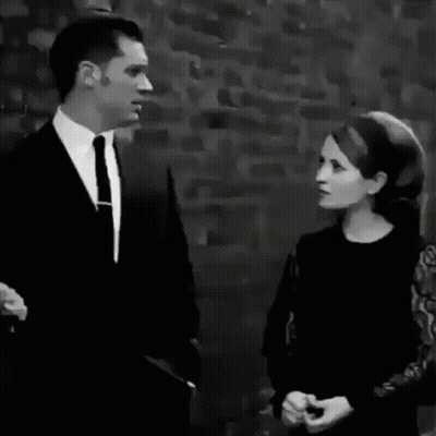 A Black And White Photo Of A Man In A Suit And Tie Standing In Front Of A Brick Wall GIF