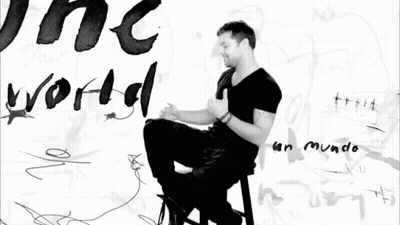 A Black And White Photo Of A Man Sitting In Front Of A Wall With The Word World Written On It GIF