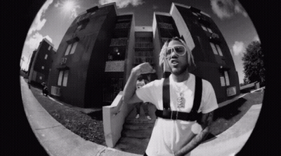 A Black And White Photo Of A Man Standing In Front Of A Building Wearing Sunglasses . GIF