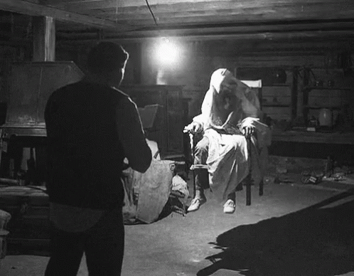 A Black And White Photo Of A Man Standing In Front Of A Ghost In A Chair GIF