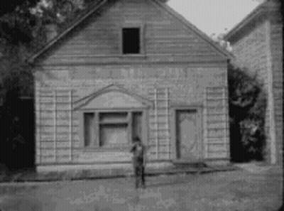 A Black And White Photo Of A Man Standing In Front Of A Small Wooden Building . GIF
