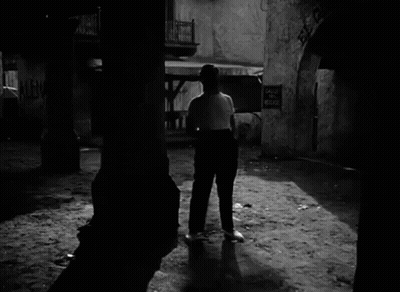 A Black And White Photo Of A Man Walking In Front Of A Sign That Says Calle Del Medio GIF