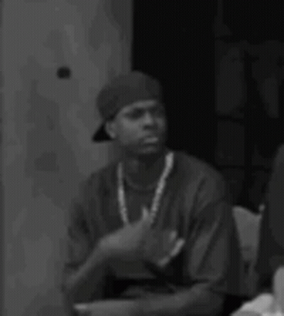 A Black And White Photo Of A Man Wearing A Hat And A Necklace Sitting Down . GIF