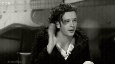 A Black And White Photo Of A Man With Curly Hair Sitting At A Table With His Hand In His Hair . GIF