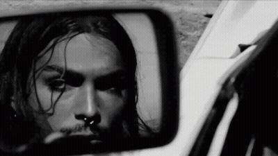 A Black And White Photo Of A Man With Long Hair And A Nose Ring Looking At Himself In A Rear View Mirror . GIF
