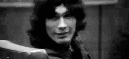 A Black And White Photo Of A Man With Long Hair Sitting In A Chair And Smiling . GIF