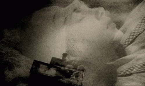A Black And White Photo Of A Person Laying On A Bed With A House In The Background . GIF