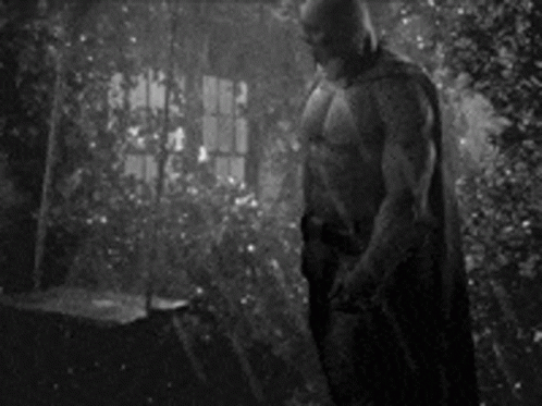 A Black And White Photo Of A Shirtless Man In A Batman Costume Standing In Front Of A Window . GIF