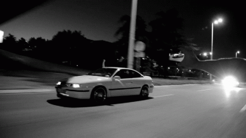 A Black And White Photo Of A White Car Driving Down A Street GIF