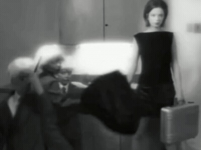 A Black And White Photo Of A Woman In A Black Dress Holding A Silver Briefcase GIF