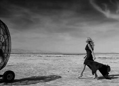A Black And White Photo Of A Woman In A Black Dress Walking In The Desert GIF