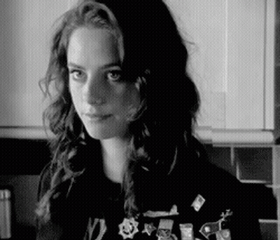A Black And White Photo Of A Woman In A Kitchen Wearing A Sweater With Pins On It . GIF