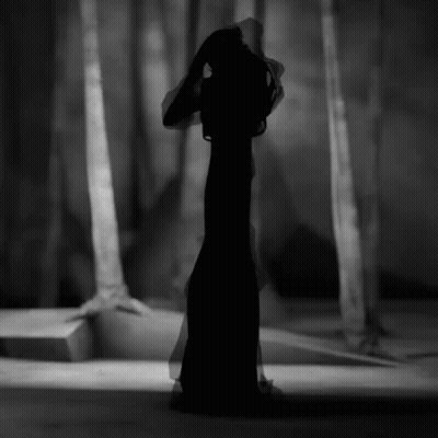 A Black And White Photo Of A Woman In A Long Black Dress In A Forest GIF