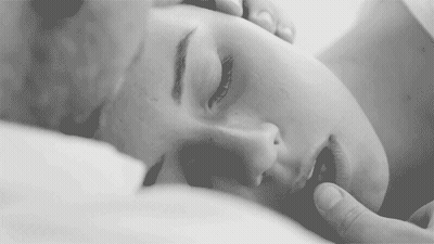 A Black And White Photo Of A Woman Laying On A Bed With Her Eyes Closed And A Man Touching Her Face . GIF