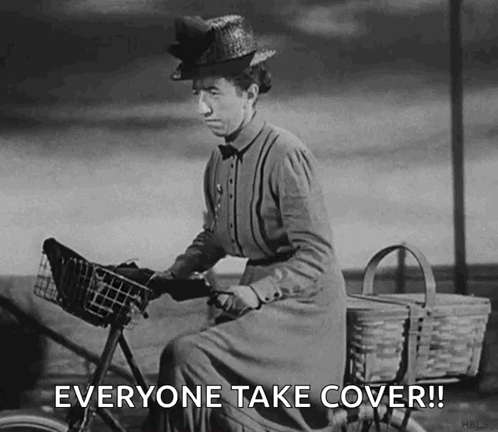 A Black And White Photo Of A Woman Riding A Bicycle With The Words Everyone Take Cover ! GIF