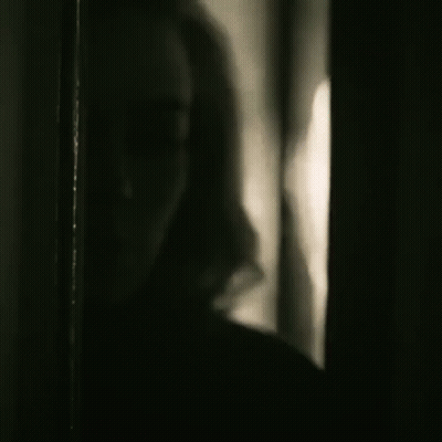 A Black And White Photo Of A Woman Standing In A Dark Room Looking Out A Window . GIF