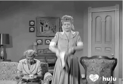 A Black And White Photo Of A Woman Standing In A Living Room Holding A Cat . GIF