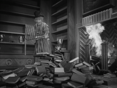 A Black And White Photo Of A Woman Standing In Front Of A Pile Of Books GIF