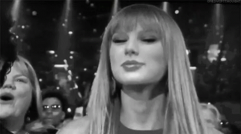 A Black And White Photo Of A Woman With Long Blonde Hair And Bangs Standing In Front Of A Crowd . GIF