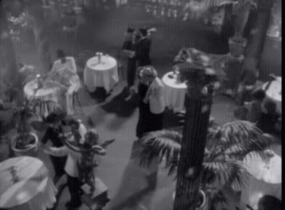 A Black And White Photo Of People Dancing In A Room With Tables GIF