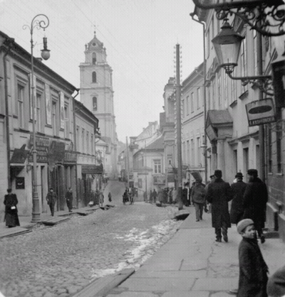 A Black And White Photo Of People Walking Down A Cobblestone Street With A Sign That Says ' Leningradska ' GIF