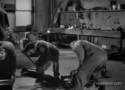 A Black And White Photo Of Two Men Working On A Car Engine In A Garage . GIF