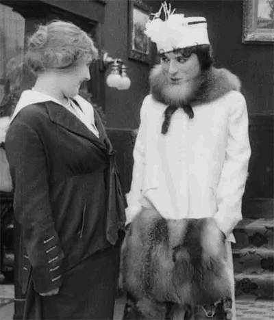 A Black And White Photo Of Two Women Talking To Each Other . One Of The Women Is Wearing A Fur Coat . GIF