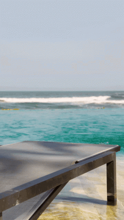 A Black Lounge Chair Sits On A Beach With The Ocean In The Background GIF