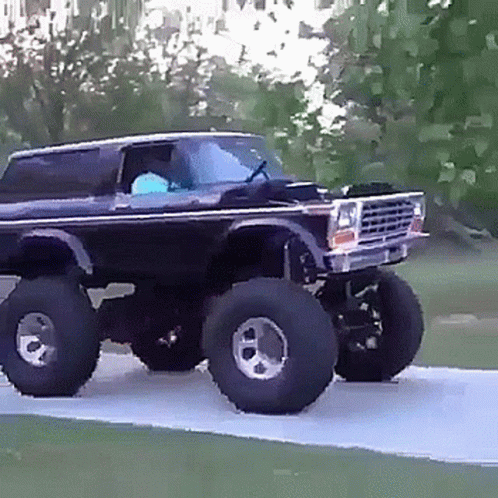 A Black Truck With Huge Tires Is Driving Down A Road . GIF