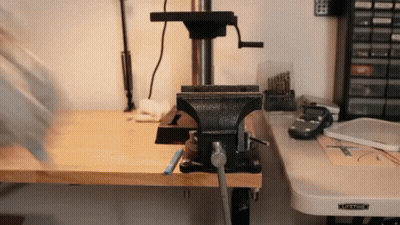 A Black Vise Sits On A Wooden Workbench Next To A Drill Press GIF