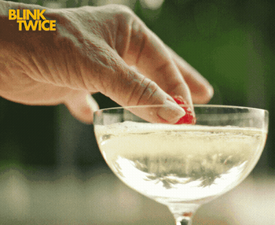 A Blink Twice Ad Shows A Person Putting A Strawberry In A Glass Of Champagne GIF