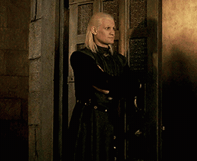 A Blonde Woman In A Black Coat Is Standing In Front Of A Door GIF