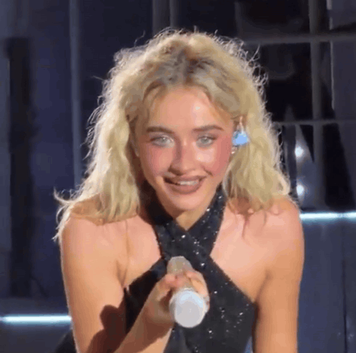 A Blonde Woman In A Black Dress Is Holding A Microphone And Smiling GIF