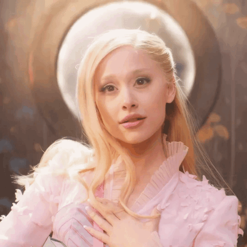 A Blonde Woman In A Pink Dress Holds Her Hands To Her Chest GIF