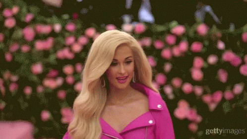 A Blonde Woman In A Pink Jacket Stands In Front Of A Wall Of Roses GIF